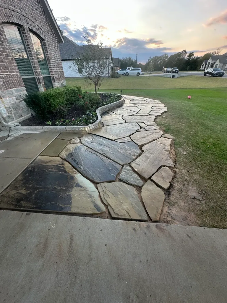 Curved flagstone front walkway beside a stone-edged flower bed with landscape lighting at sunset