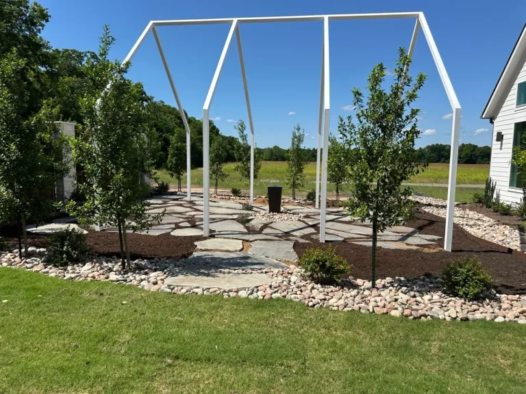 Flagstone patio under a white pergola frame, edged with river rock and mulch beds with newly planted trees
